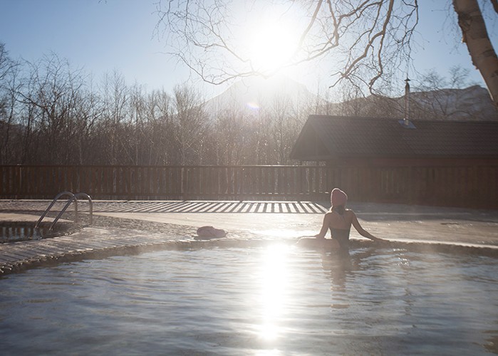 Woman relaxing in a steaming outdoor thermal pool on a bright winter morning with bare trees and a wooden cabin behind